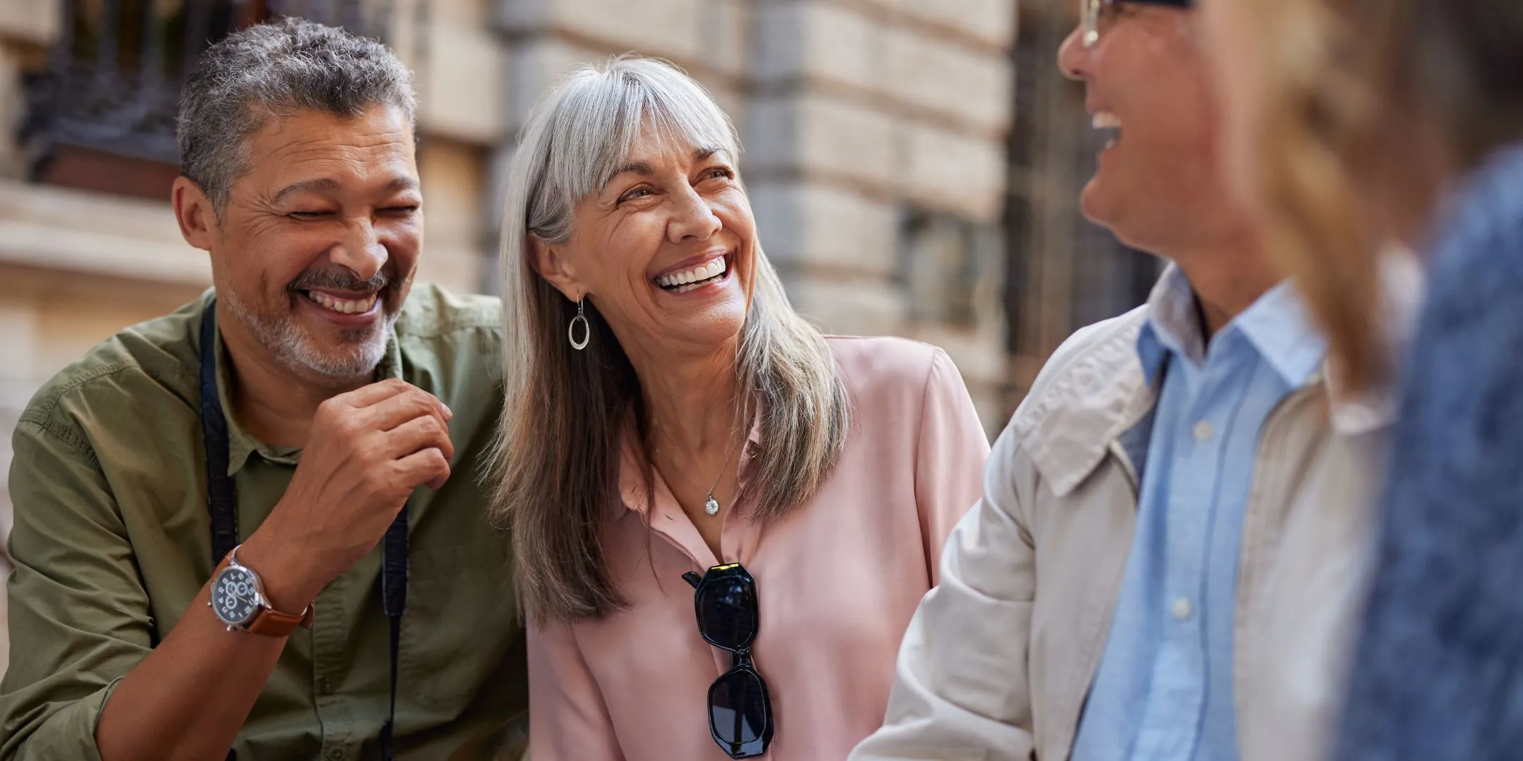 A group of four older adults stand close together outdoors, laughing warmly. A man in an olive shirt and woman in a pink blouse share a laugh in the foreground, with two more people smiling beside them. A pale stone building is blurred in the background