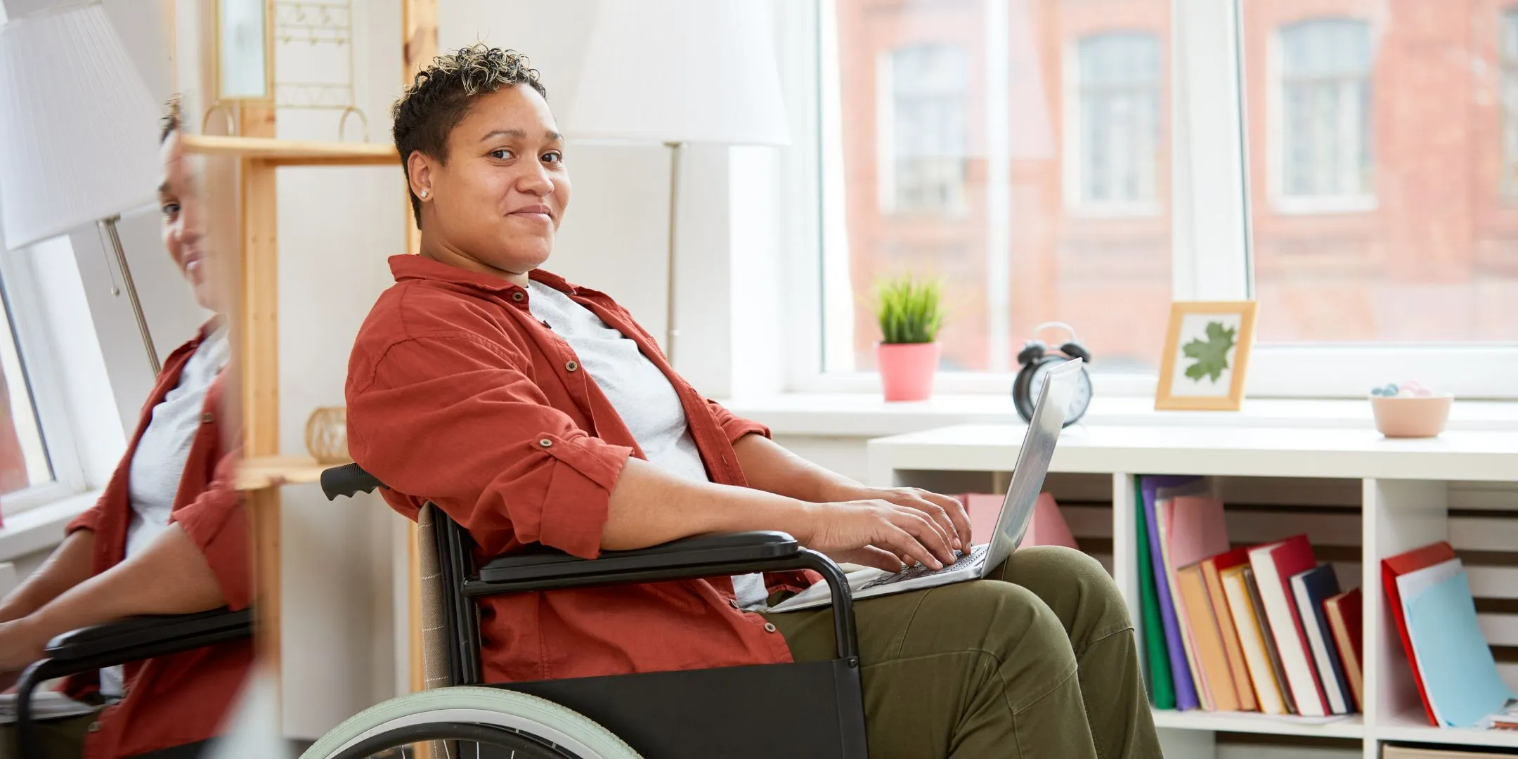 A person sits in a wheelchair at a desk, using a laptop in a bright, comfortable room. They appear relaxed and focused.