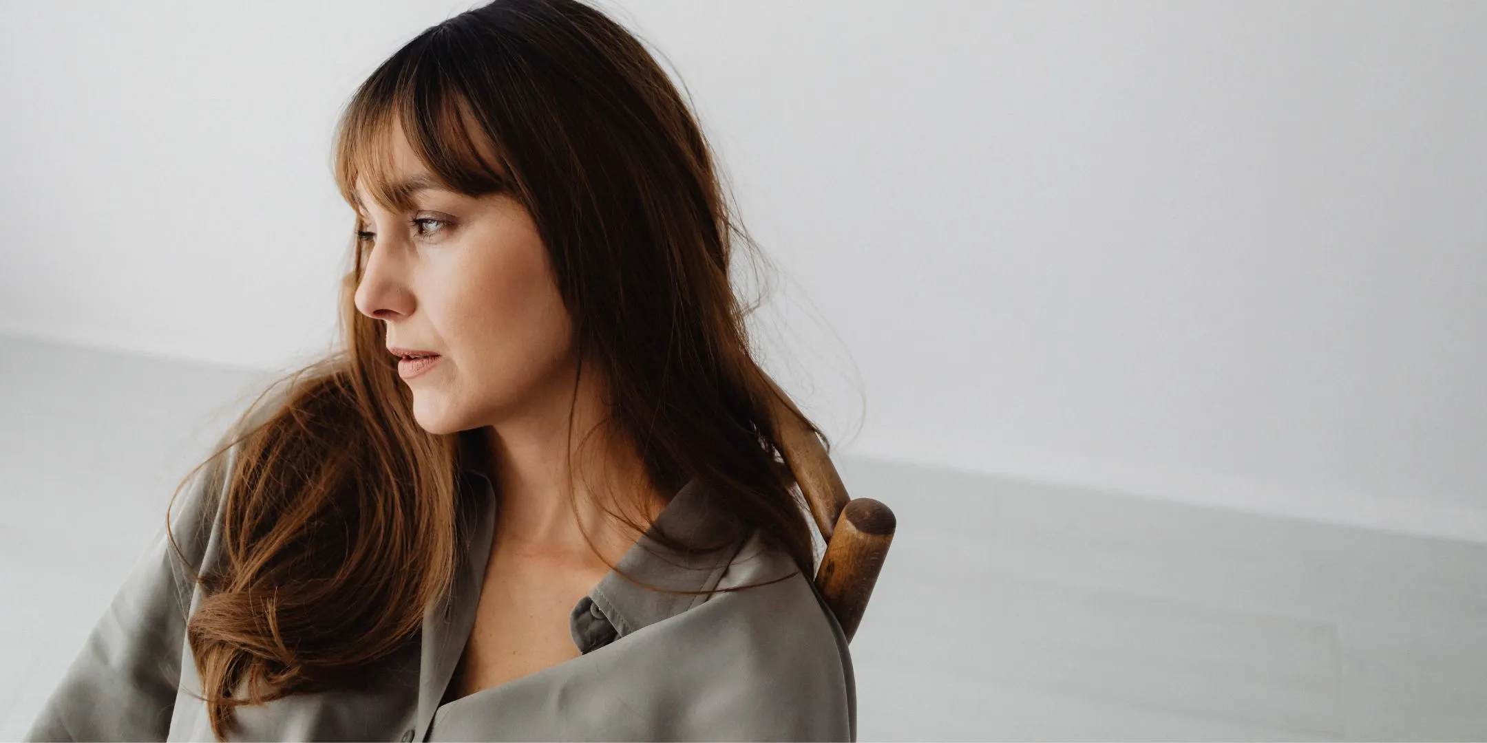 The image shows a woman seated indoors against a simple, light-colored background. She’s turned slightly to the side, looking off to her left rather than at the camera. Her expression appears thoughtful and a bit serious, as if she’s reflecting on something or listening intently.  She has long brown hair with soft bangs and is wearing a muted, grey blouse. The setting is minimal and uncluttered, which draws attention to her face and posture. A wooden chair back is visible behind her, adding a subtle, natural element to the scene.