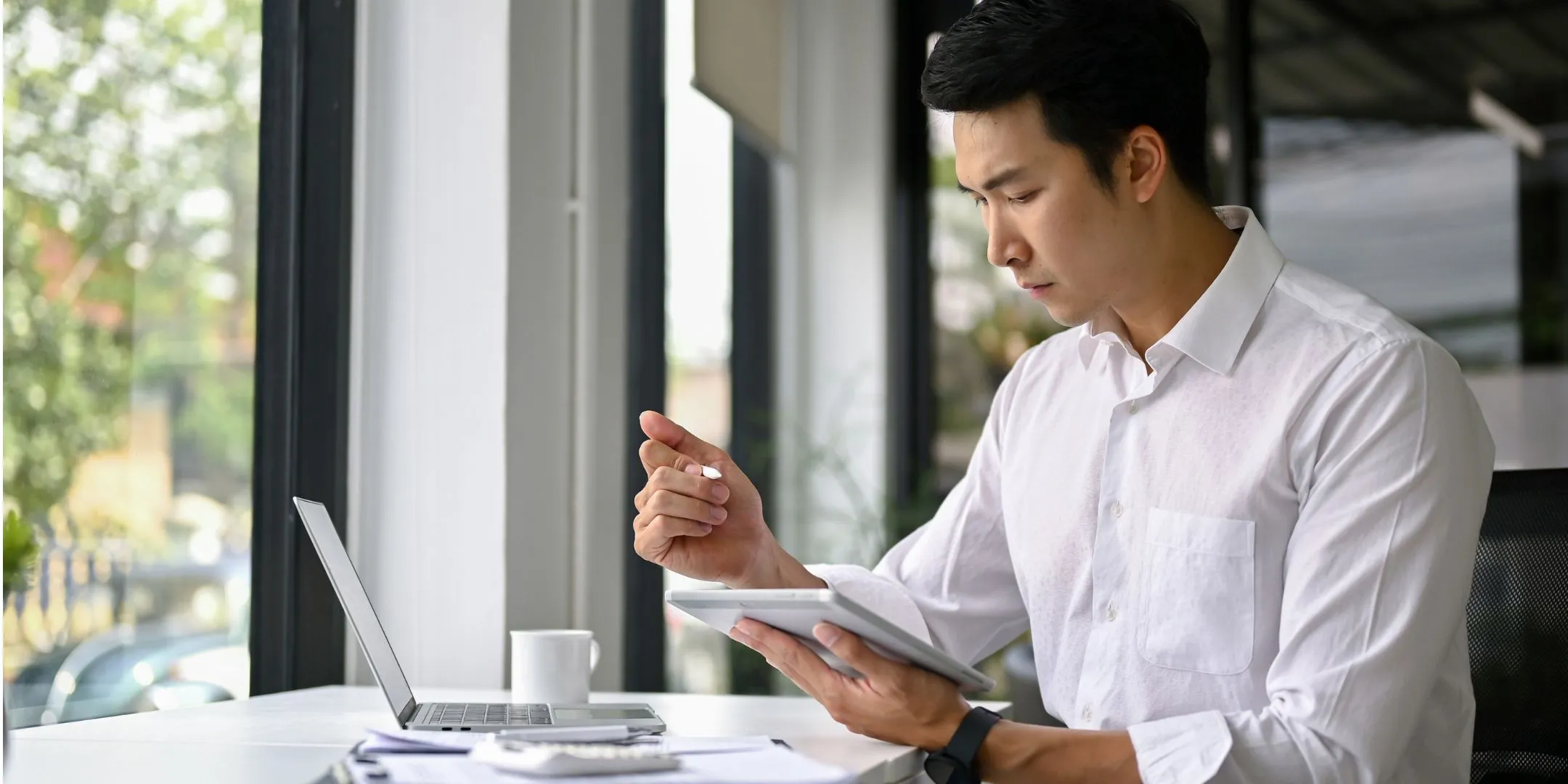 This image shows a man sitting at a desk near a large window, working with both a laptop and a tablet. He’s holding a stylus and looking down at the tablet with a focused, thoughtful expression, as if reviewing information or doing calculations.  He’s dressed in a white shirt, giving a professional or work-related feel. On the desk, there’s also a cup and some papers, suggesting a typical workspace setup. The natural light from the window creates a calm, productive atmosphere.  Overall, the scene conveys concentration, problem-solving, and focused work.