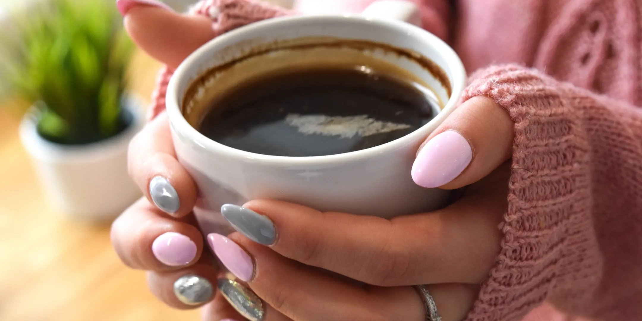 A close-up of hands holding a cup of coffee. The person has neatly painted nails and is wearing a soft sweater, creating a calm, cozy atmosphere.