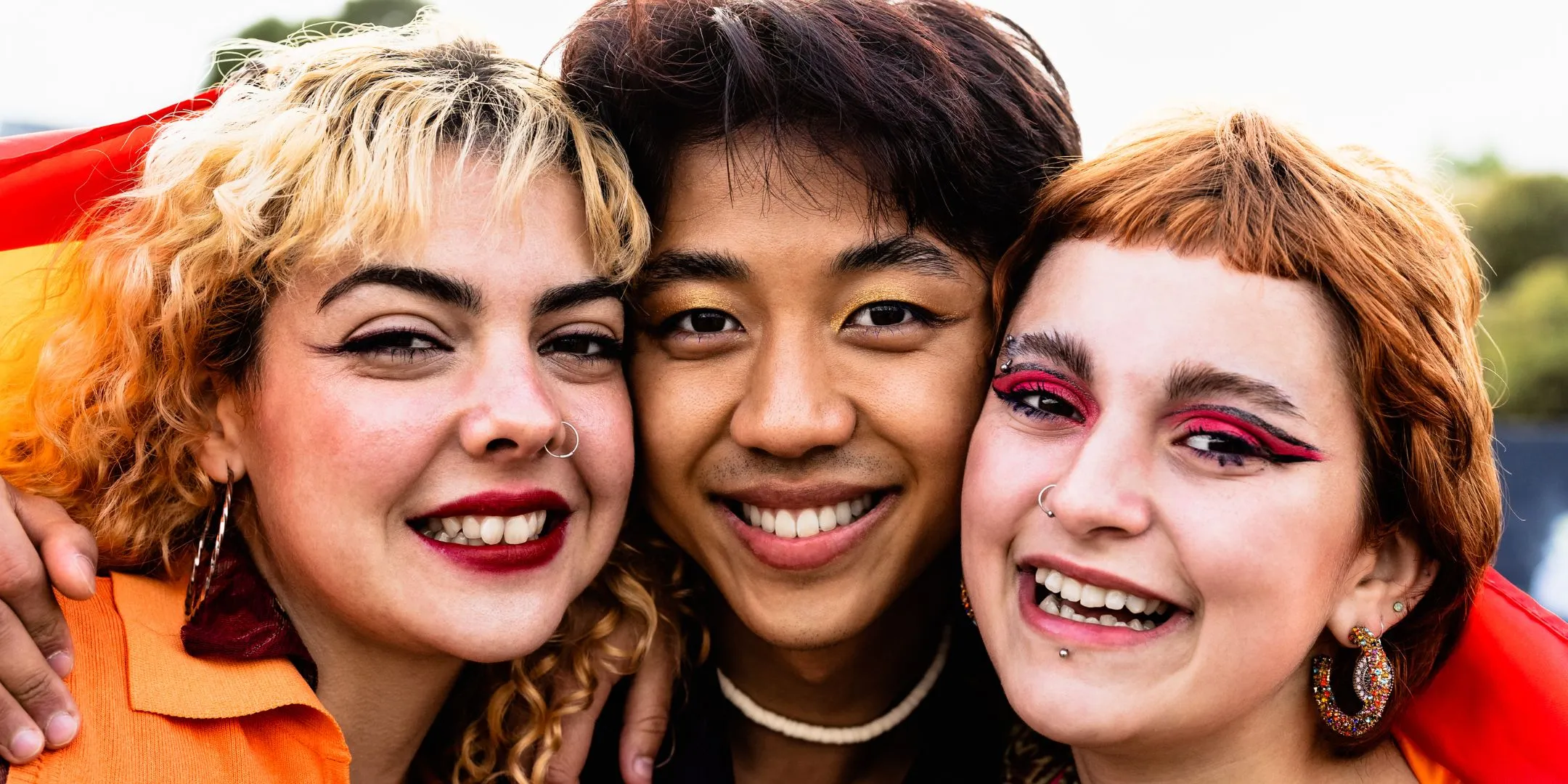 Three friends pose closely together outdoors, smiling at the camera in colourful clothing and expressive makeup.