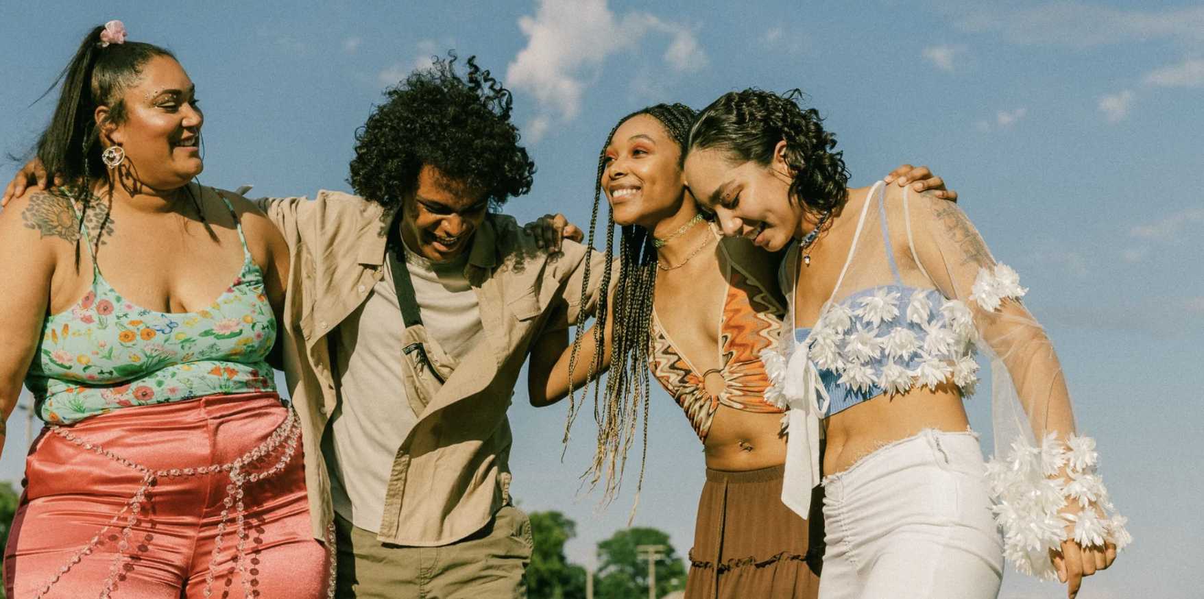 Four people walking outdoors with arms around each other, smiling and laughing, dressed in bright festival-style clothing against a blue sky.