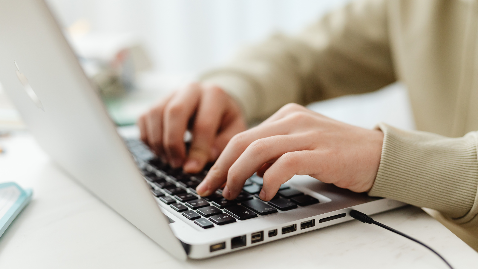 Close-up of hands typing on a laptop keyboard, with a bright, softly blurred room in the background.