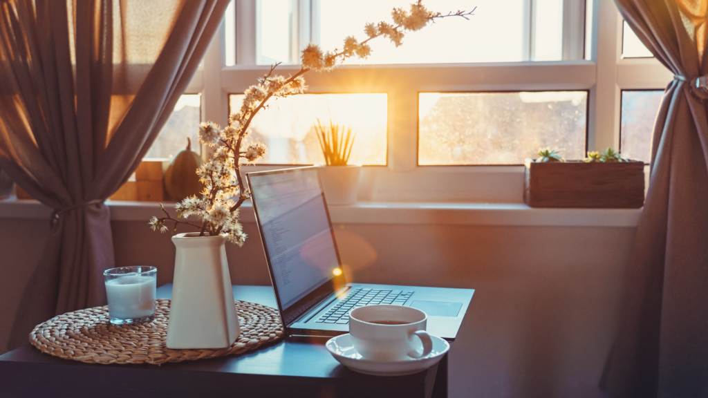Laptop, cup of tea, and a vase of blossom on a dark wood desk beside a sunlit window, curtains open to golden evening light. Image from Canva
