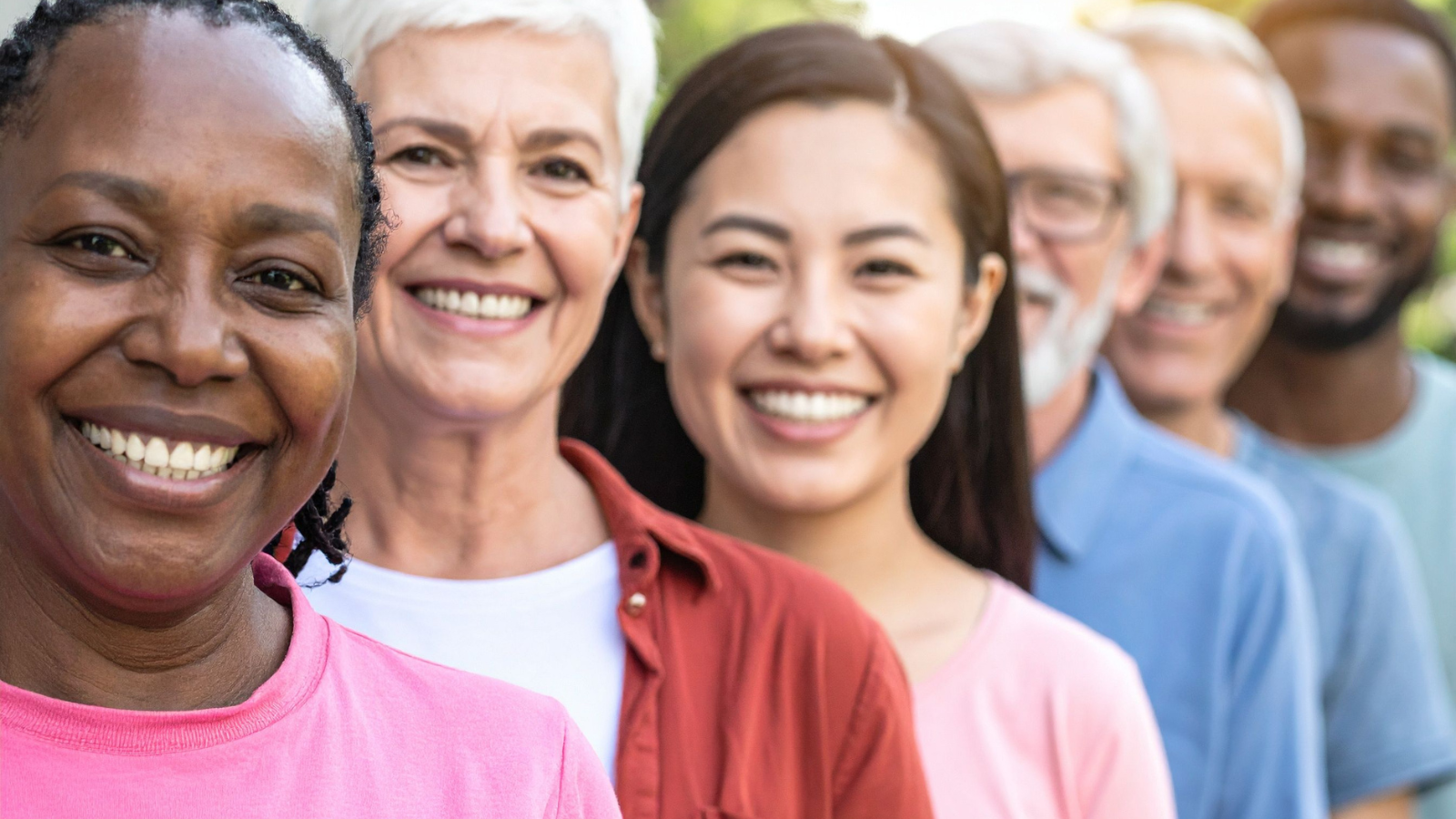 Row of people of different ages and races standing in a line outdoors, all smiling at the camera.