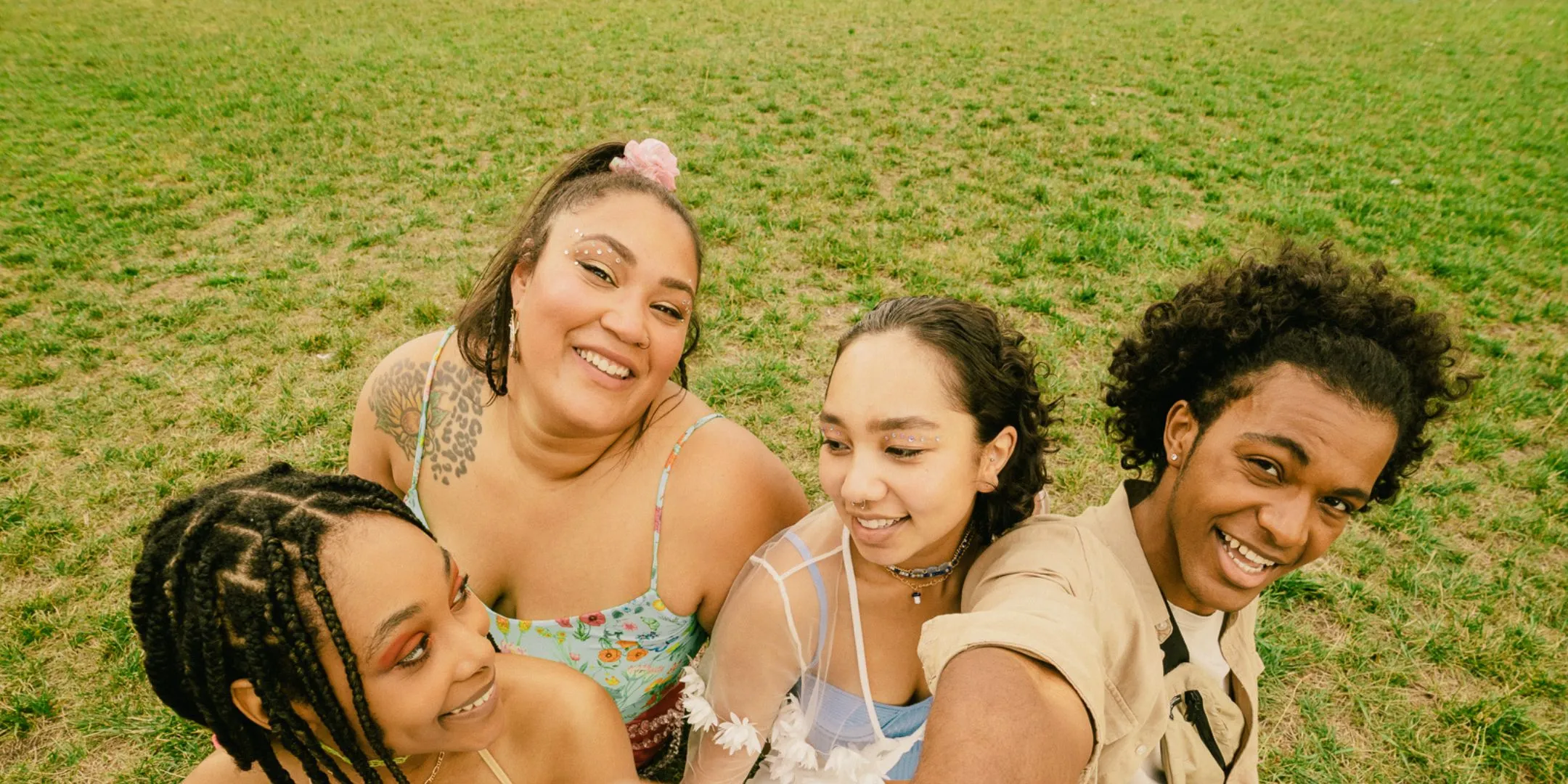Four friends sit together on a grassy field, smiling and enjoying time together outdoors.