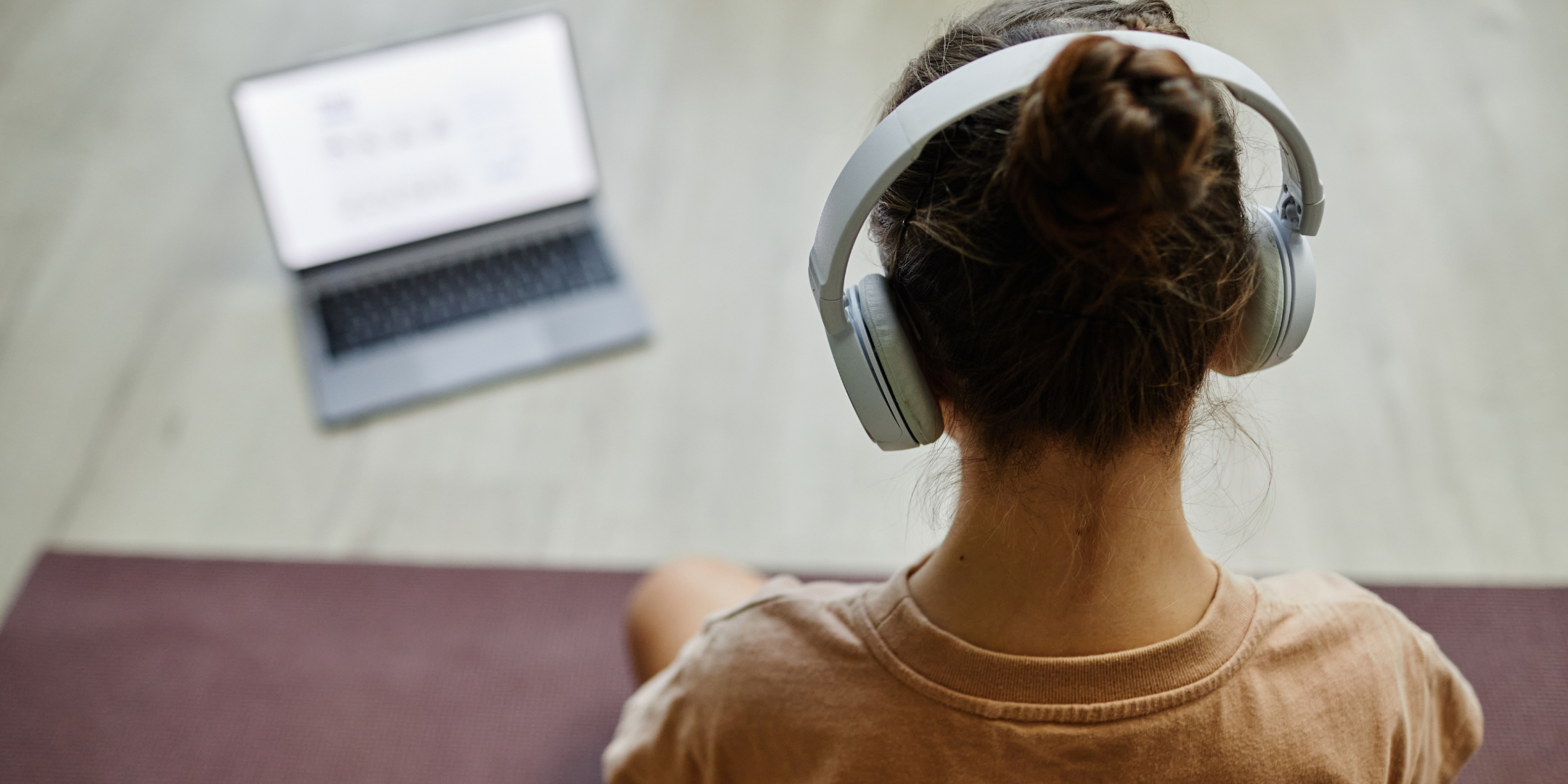 Person wearing over-ear headphones, seated on a mat and facing an open laptop in a calm indoor setting.